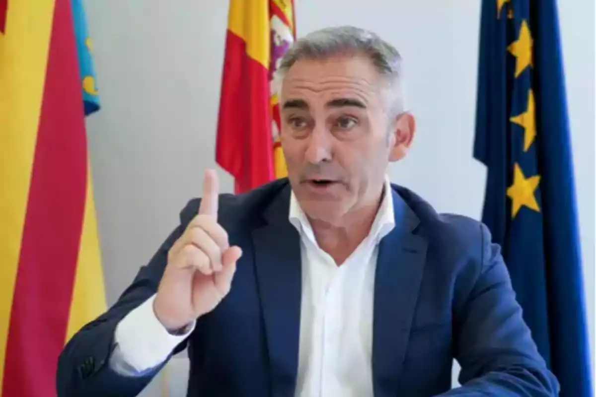 Hombre de cabello canoso con traje azul y camisa blanca levantando el dedo índice mientras habla, sentado frente a banderas oficiales Hombre de cabello canoso con traje azul y camisa blanca levantando el dedo índice mientras habla, sentado frente a banderas oficiales