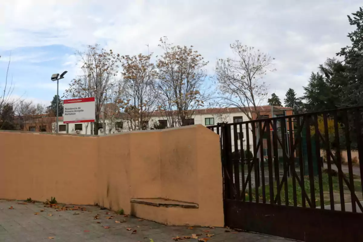 Entrada con portón metálico y muro color beige, al fondo se observan árboles sin hojas y un edificio blanco con un letrero que indica Residencia de Primera Acogida Hortaleza Entrada con portón metálico y muro color beige, al fondo se observan árboles sin hojas y un edificio blanco con un letrero que indica Residencia de Primera Acogida Hortaleza