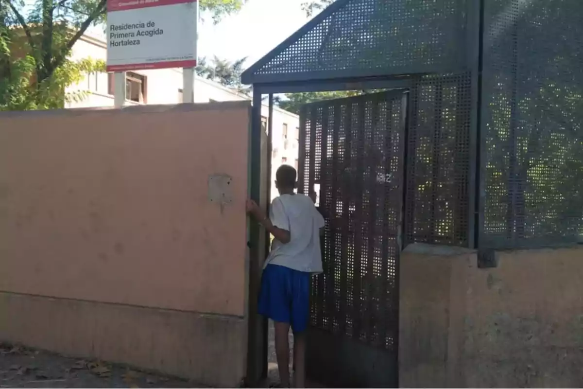 Un joven con camiseta blanca y pantalones cortos azules entra por una puerta metálica en una residencia de primera acogida, junto a un muro y un cartel identificativo. Un joven con camiseta blanca y pantalones cortos azules entra por una puerta metálica en una residencia de primera acogida, junto a un muro y un cartel identificativo.