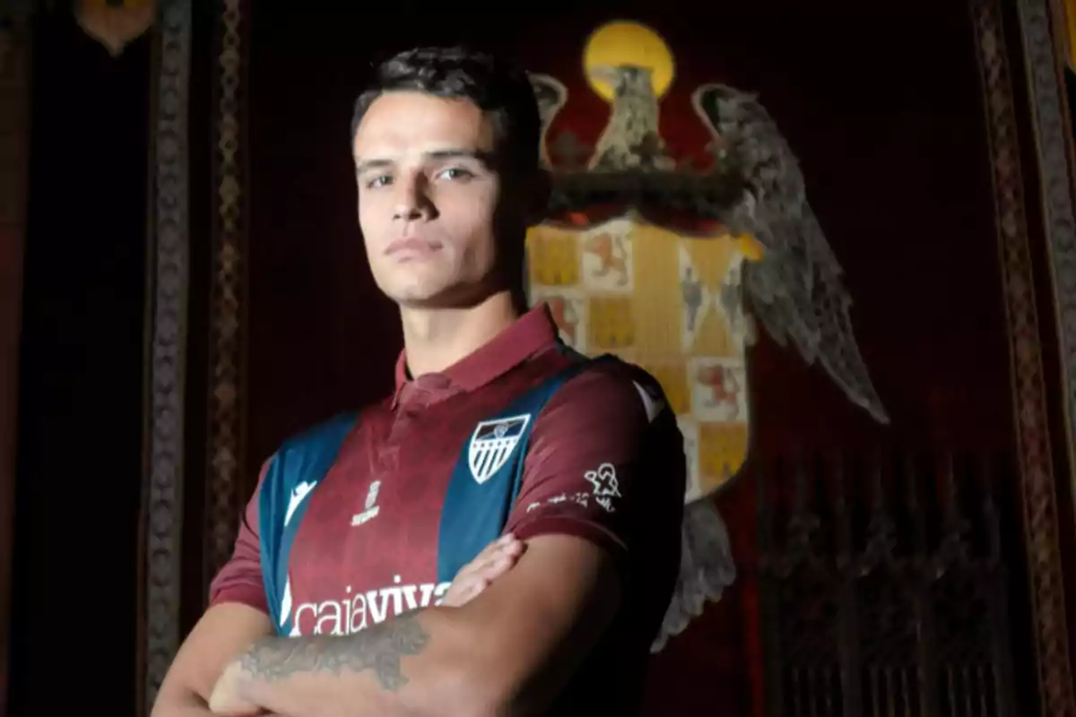 Jugador de fútbol con uniforme azul y burdeos posando con los brazos cruzados frente a un escudo grande en el fondo Jugador de fútbol con uniforme azul y burdeos posando con los brazos cruzados frente a un escudo grande en el fondo