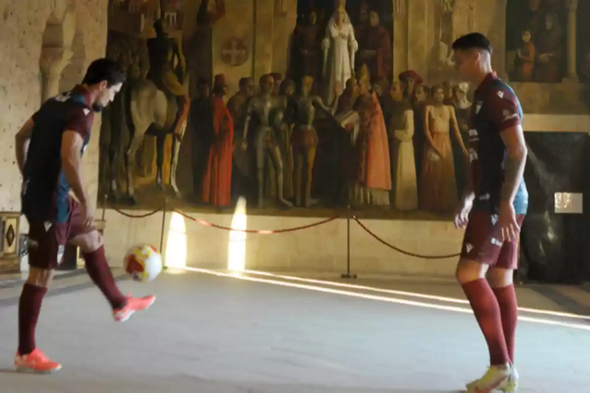 Dos futbolistas con uniforme granate juegan con un balón dentro de una sala antigua decorada con un gran mural histórico al fondo Dos futbolistas con uniforme granate juegan con un balón dentro de una sala antigua decorada con un gran mural histórico al fondo
