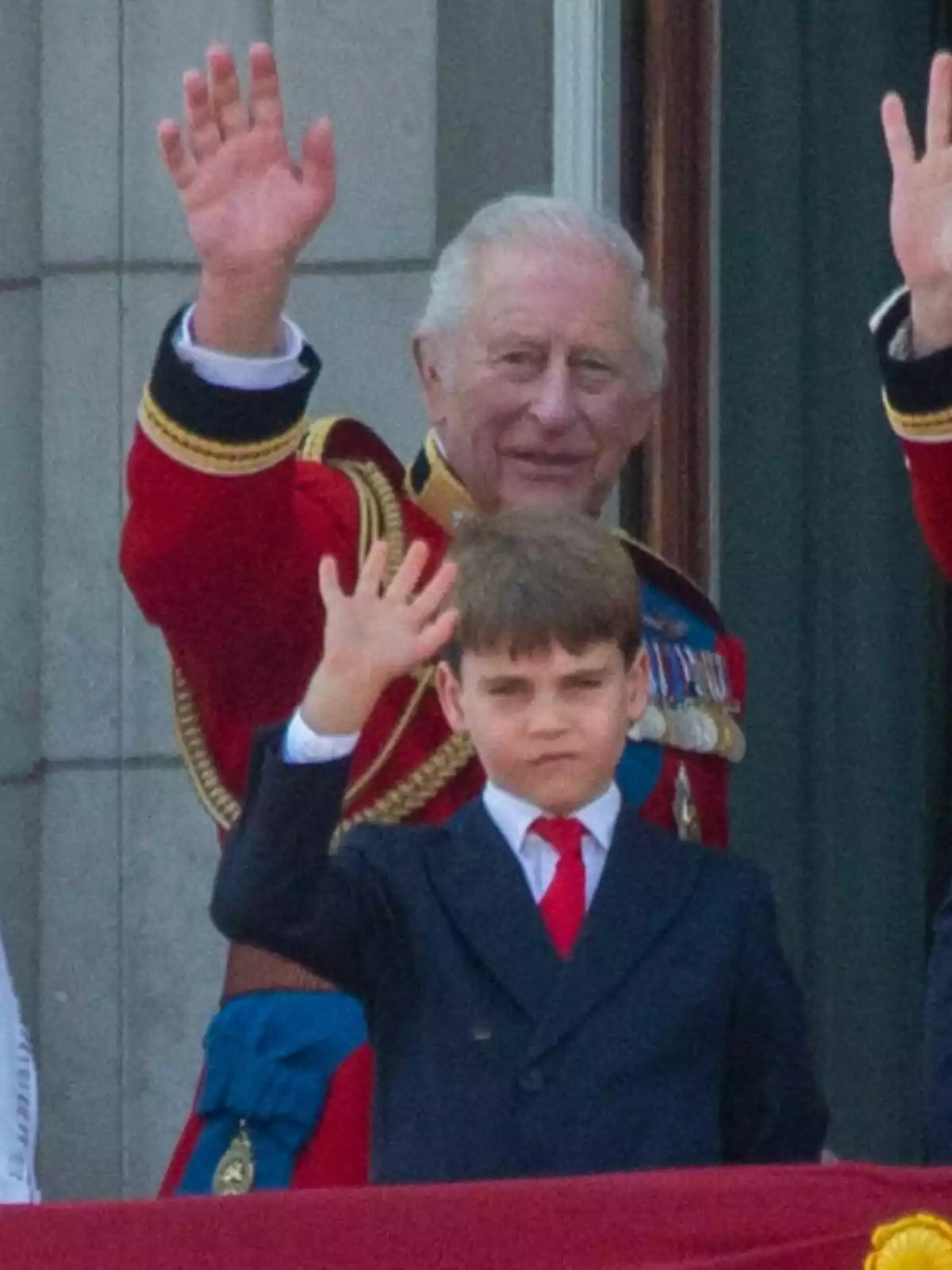Carlos III con uniforme rojo y el príncipe Louis con traje oscuro y corbata roja saludan desde un balcón Carlos III con uniforme rojo y el príncipe Louis con traje oscuro y corbata roja saludan desde un balcón