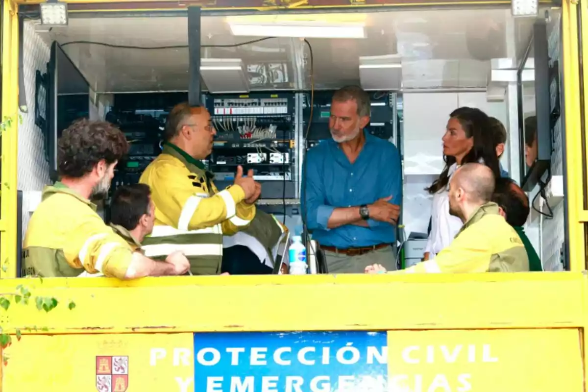 Several people are talking inside a civil protection and emergency mobile unit; some are wearing yellow uniforms and others are in civilian clothes, surrounded by electronic equipment. Several people are talking inside a civil protection and emergency mobile unit; some are wearing yellow uniforms and others are in civilian clothes, surrounded by electronic equipment.