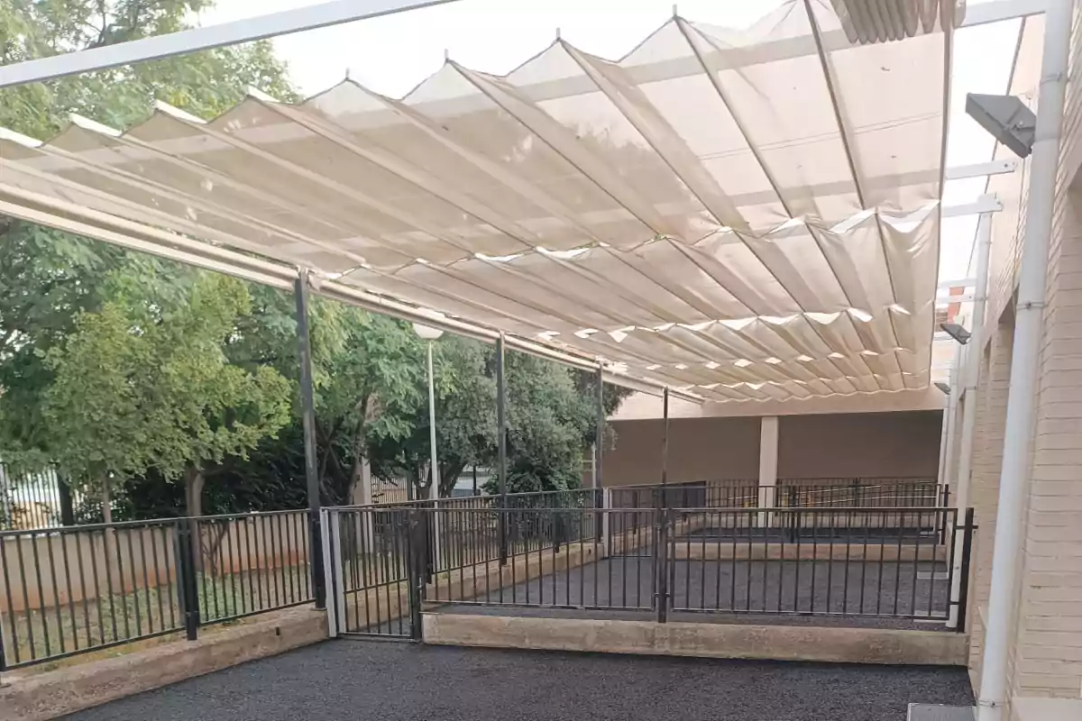 Estructura metálica con techo de lona blanca plegable sobre un patio cercado con barandales negros y árboles al fondo Estructura metálica con techo de lona blanca plegable sobre un patio cercado con barandales negros y árboles al fondo