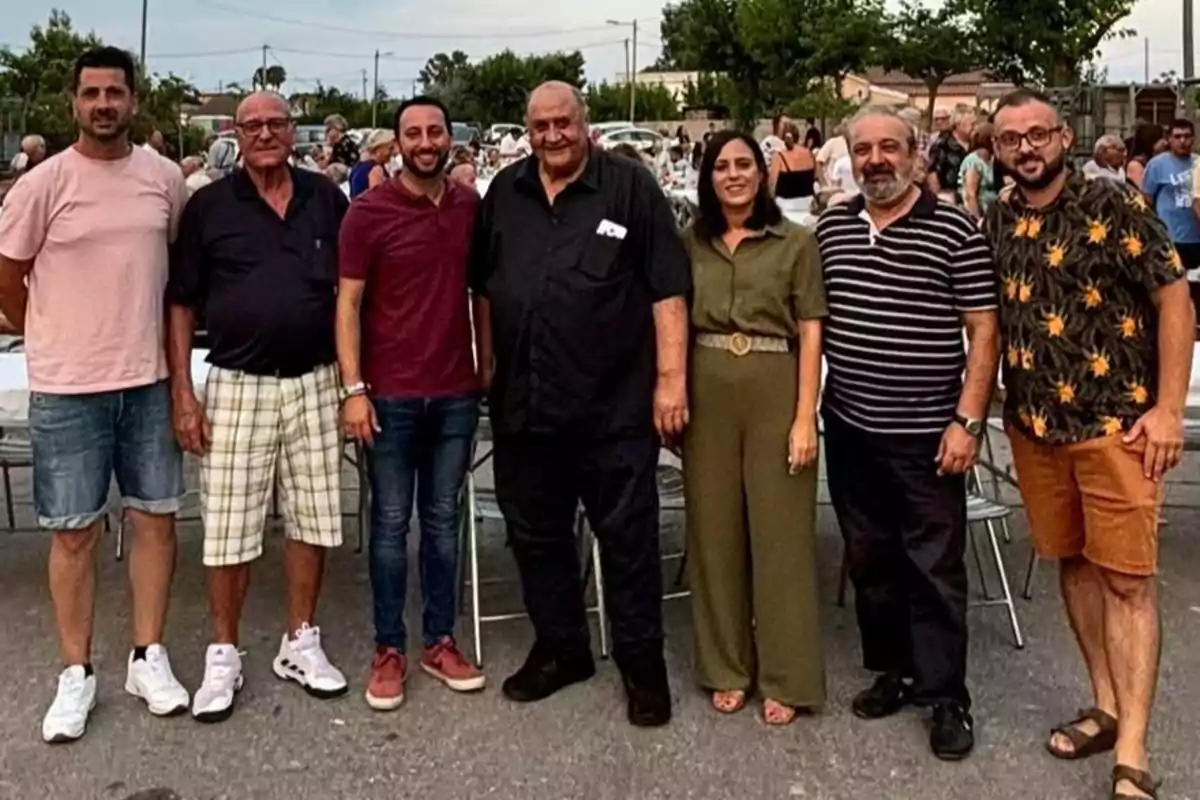 Siete personas posan sonrientes al aire libre en un evento social con mesas y gente al fondo Siete personas posan sonrientes al aire libre en un evento social con mesas y gente al fondo