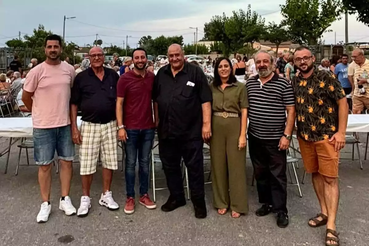 Siete personas posan sonrientes al aire libre durante un evento social con mesas y sillas al fondo y más gente reunida en un ambiente relajado