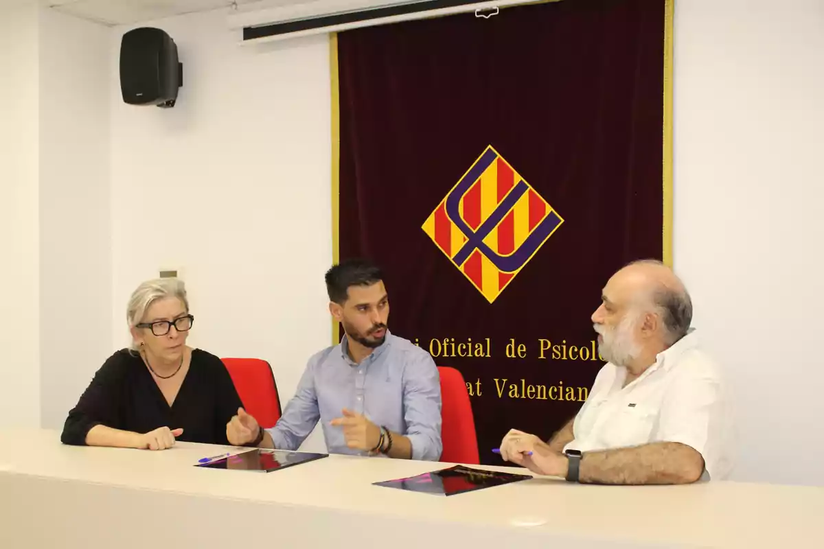 Tres personas sentadas en una mesa conversando frente a un estandarte con un logotipo y letras doradas Tres personas sentadas en una mesa conversando frente a un estandarte con un logotipo y letras doradas