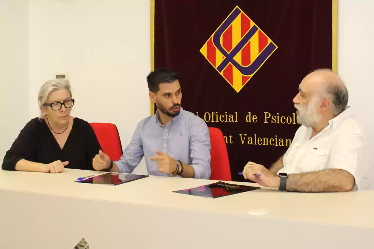 Tres personas sentadas en una mesa conversan en una sala con un cartel del Colegio Oficial de Psicología de la Comunitat Valenciana al fondo