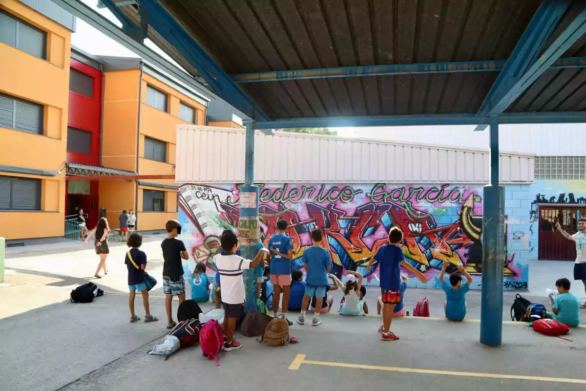 Un grupo de niños y niñas con mochilas se reúne en el patio de una escuela junto a una pared decorada con un colorido mural de graffiti, mientras algunos adultos caminan cerca del edificio escolar de fondo