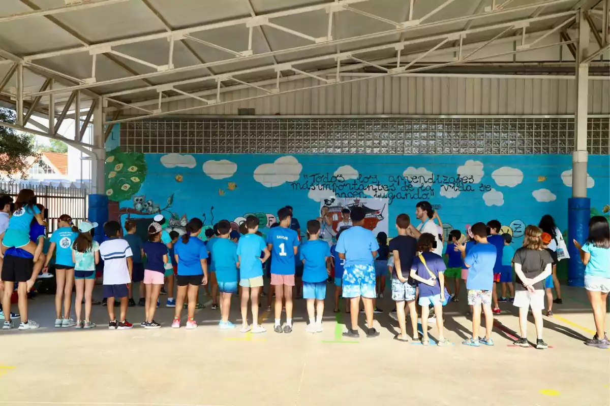 Un grupo de niños y adultos se encuentra reunido bajo un techo en un patio escolar, todos de espaldas, observando a unas personas frente a un mural colorido con nubes y un mensaje escrito en la pared. Un grupo de niños y adultos se encuentra reunido bajo un techo en un patio escolar, todos de espaldas, observando a unas personas frente a un mural colorido con nubes y un mensaje escrito en la pared.