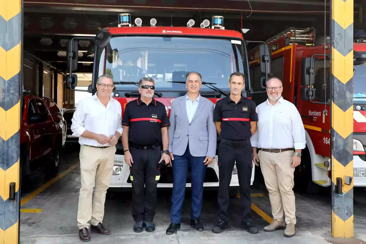 Cinco hombres posan frente a un camión de bomberos dentro de una estación, dos de ellos llevan uniforme de bombero y los otros tres visten ropa formal.