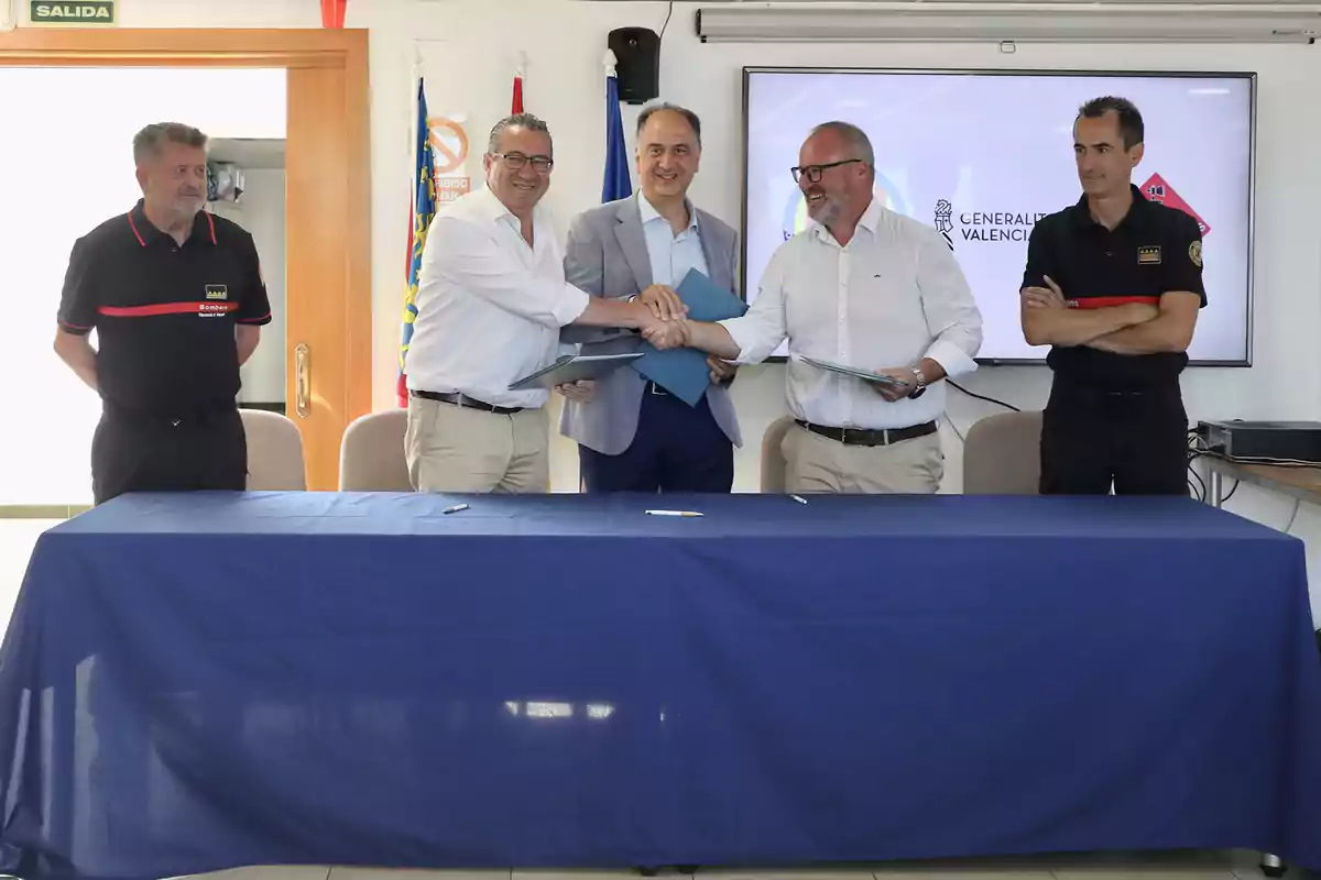 Cinco hombres posan en una sala de reuniones, tres de ellos se dan la mano sonrientes frente a una mesa cubierta con un mantel azul, mientras los otros dos observan a los lados, al fondo hay banderas y una pantalla con el logo de la Generalitat Valenciana. Cinco hombres posan en una sala de reuniones, tres de ellos se dan la mano sonrientes frente a una mesa cubierta con un mantel azul, mientras los otros dos observan a los lados, al fondo hay banderas y una pantalla con el logo de la Generalitat Valenciana.