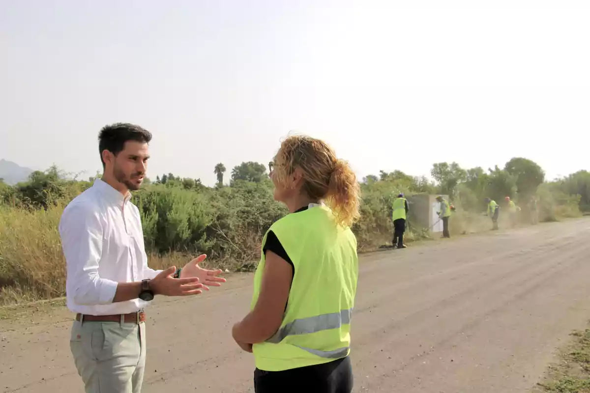 Un hombre con camisa blanca conversa con una mujer con chaleco reflectante amarillo mientras varias personas trabajan limpiando la maleza al borde de un camino rural. Un hombre con camisa blanca conversa con una mujer con chaleco reflectante amarillo mientras varias personas trabajan limpiando la maleza al borde de un camino rural.