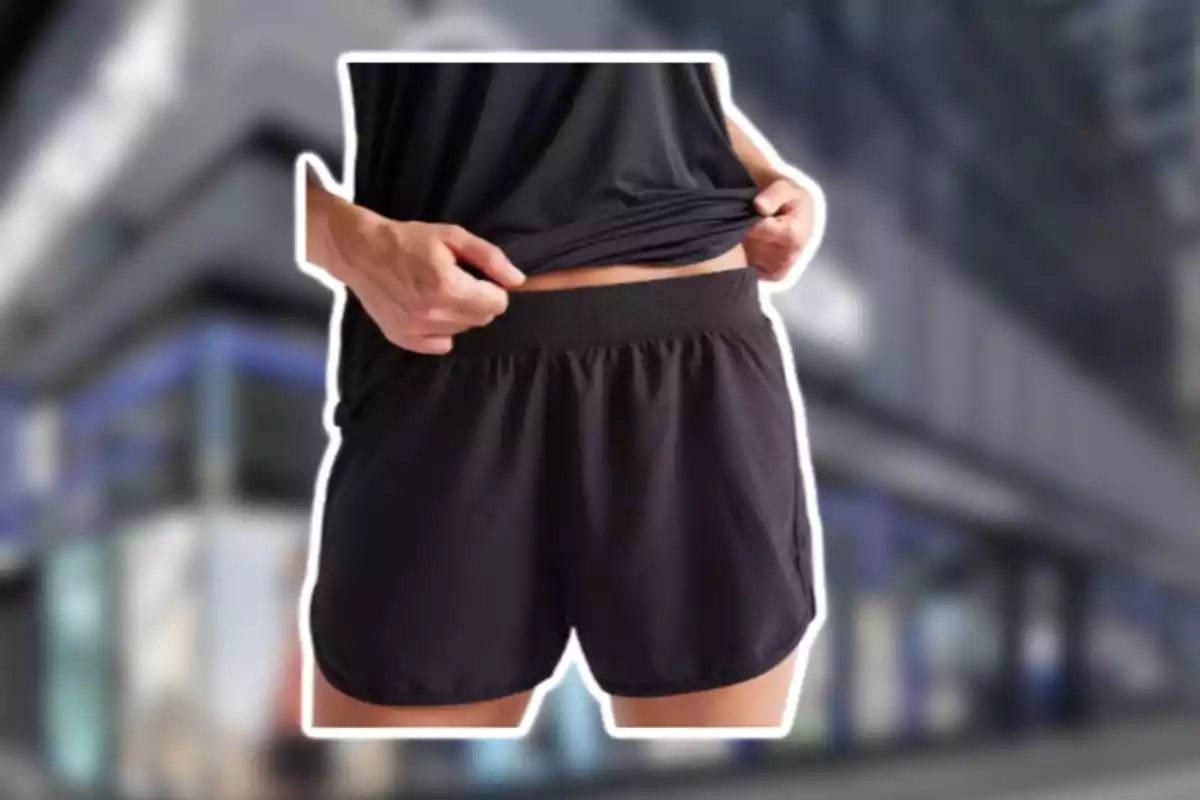 Persona usando shorts deportivos negros y levantando ligeramente la camiseta frente a un fondo urbano desenfocado Persona usando shorts deportivos negros y levantando ligeramente la camiseta frente a un fondo urbano desenfocado