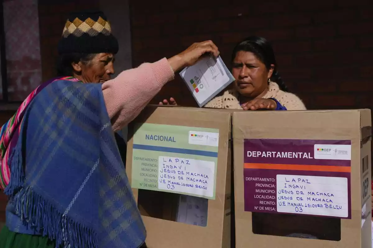 Mujer con ropa tradicional deposita su voto en una urna electoral mientras otra mujer observa durante una jornada de elecciones en Bolivia