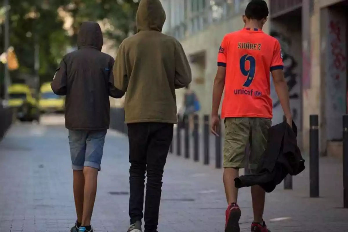 Tres adolescentes caminando por una calle urbana, dos con sudaderas con capucha y uno con camiseta deportiva roja y pantalones cortos Tres adolescentes caminando por una calle urbana, dos con sudaderas con capucha y uno con camiseta deportiva roja y pantalones cortos