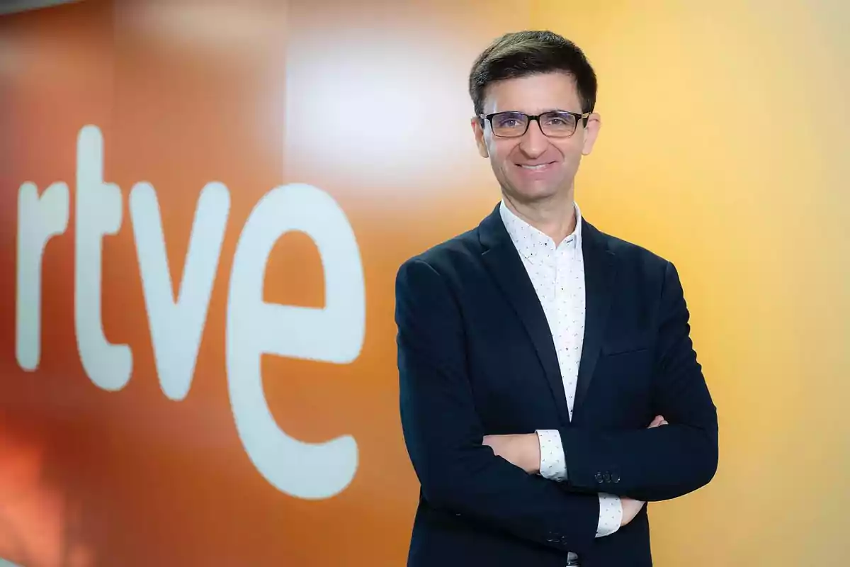 Hombre con gafas y traje oscuro sonríe con los brazos cruzados frente a una pared naranja con el logo de rtve Hombre con gafas y traje oscuro sonríe con los brazos cruzados frente a una pared naranja con el logo de rtve