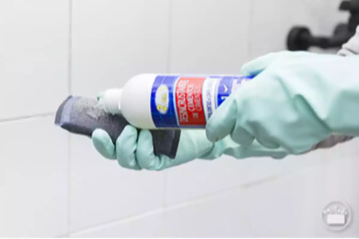 Persona con guantes verdes aplicando limpiador líquido a una esponja frente a una pared de azulejos blancos Persona con guantes verdes aplicando limpiador líquido a una esponja frente a una pared de azulejos blancos