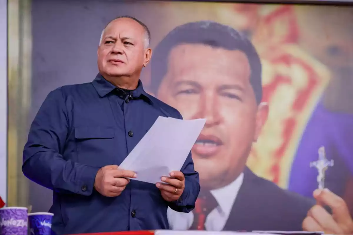 Hombre de cabello canoso con chaqueta azul sostiene unos papeles mientras está de pie frente a una imagen de otra persona en el fondo Hombre de cabello canoso con chaqueta azul sostiene unos papeles mientras está de pie frente a una imagen de otra persona en el fondo