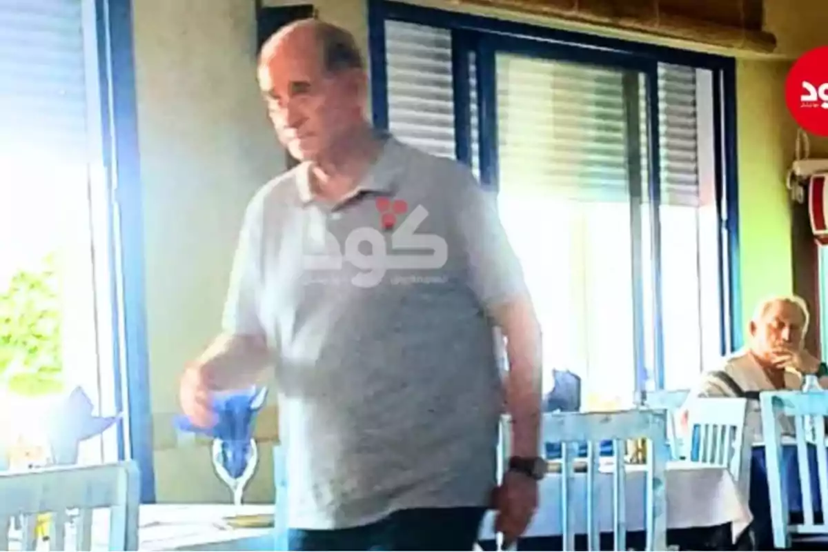 Hombre mayor de pie con camiseta gris en el interior de un restaurante iluminado, mientras otra persona está sentada al fondo