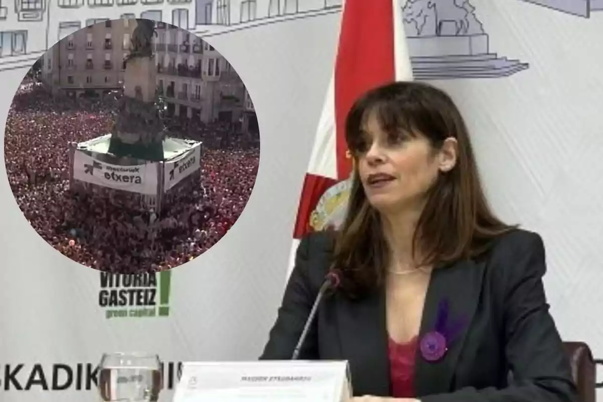 Una mujer con chaqueta oscura y blusa roja habla en una rueda de prensa con una bandera y un cartel de Vitoria Gasteiz detrás, mientras en la esquina superior izquierda aparece una imagen circular de una multitud reunida alrededor de un monumento en una plaza