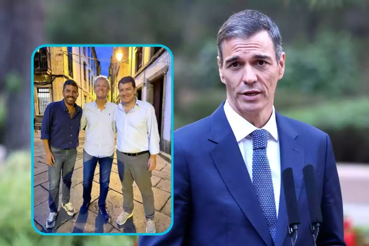 Un hombre con traje azul y corbata habla frente a dos micrófonos, mientras en la esquina izquierda aparece una foto de tres hombres sonrientes posando juntos en una calle empedrada al atardecer.