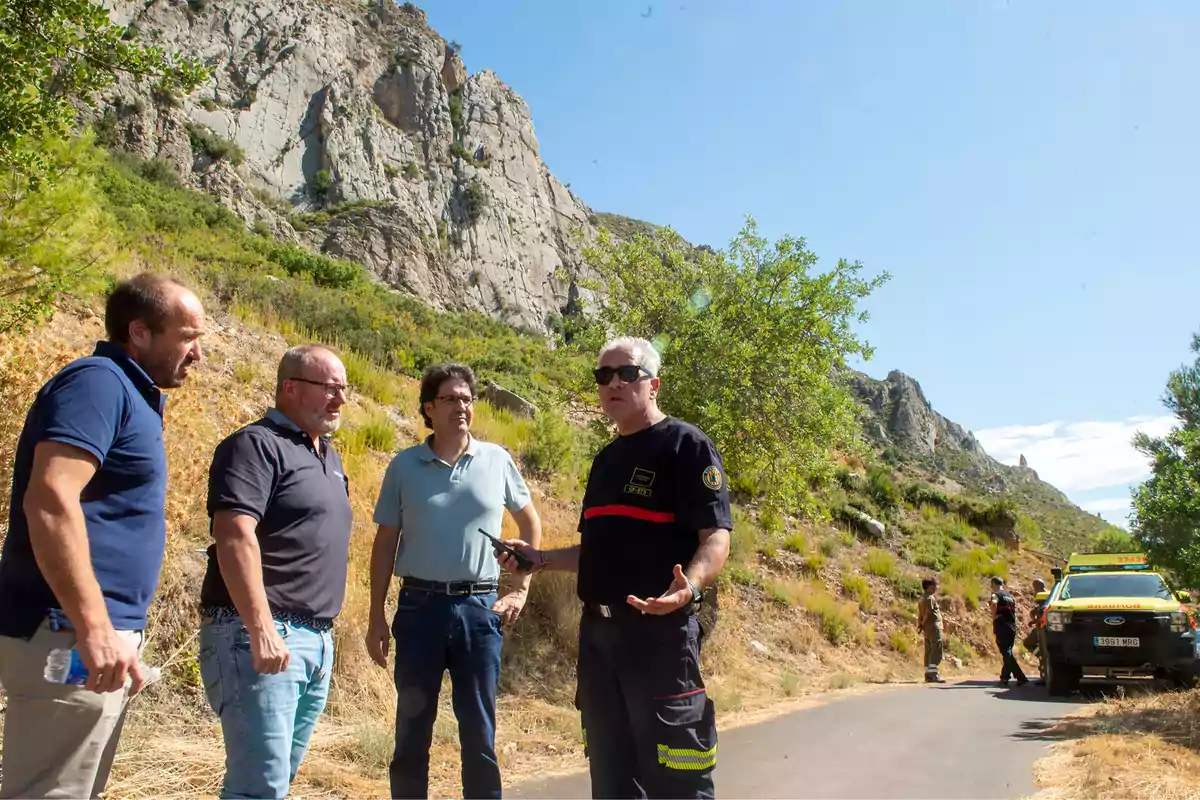 Cuatro hombres conversan en una carretera rural junto a una montaña rocosa y un vehículo de emergencia en un día soleado Cuatro hombres conversan en una carretera rural junto a una montaña rocosa y un vehículo de emergencia en un día soleado