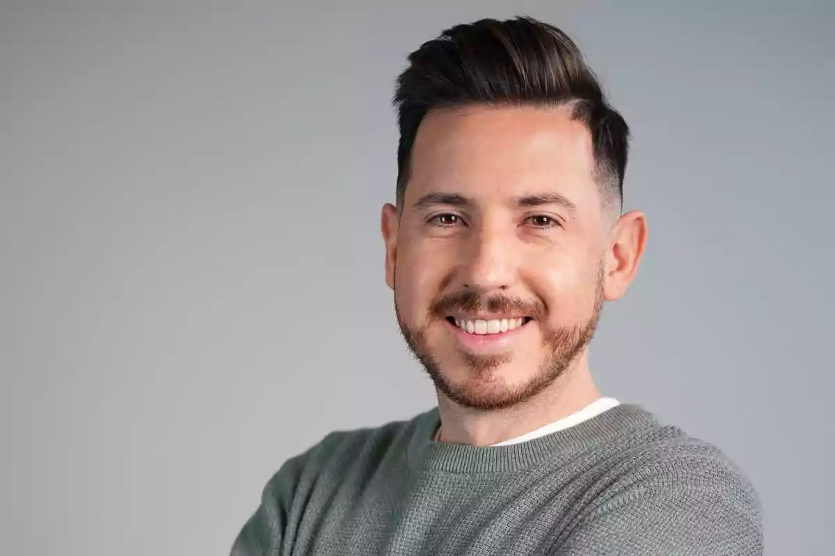 Hombre joven con barba corta y cabello oscuro peinado hacia arriba sonriendo y vistiendo un suéter gris claro sobre fondo gris