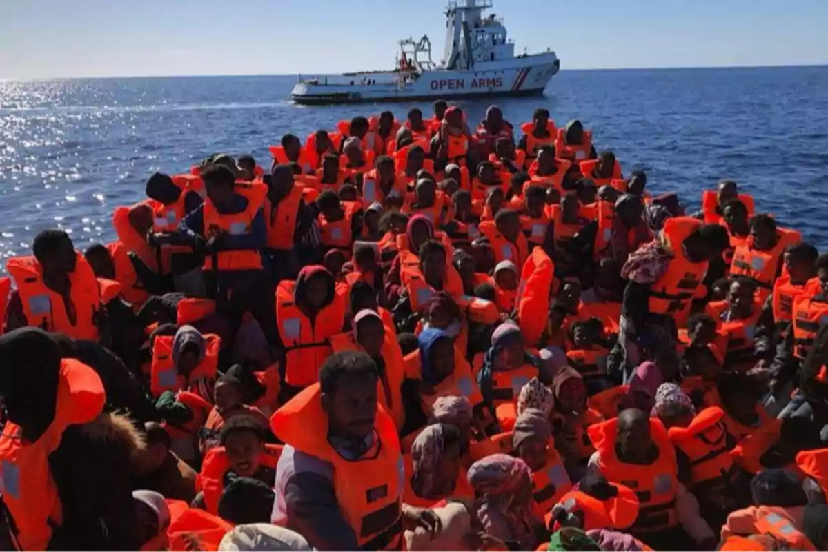 Un grupo numeroso de personas con chalecos salvavidas naranjas viaja en una embarcación en el mar mientras un barco de rescate de la organización Open Arms se encuentra cerca Un grupo numeroso de personas con chalecos salvavidas naranjas viaja en una embarcación en el mar mientras un barco de rescate de la organización Open Arms se encuentra cerca