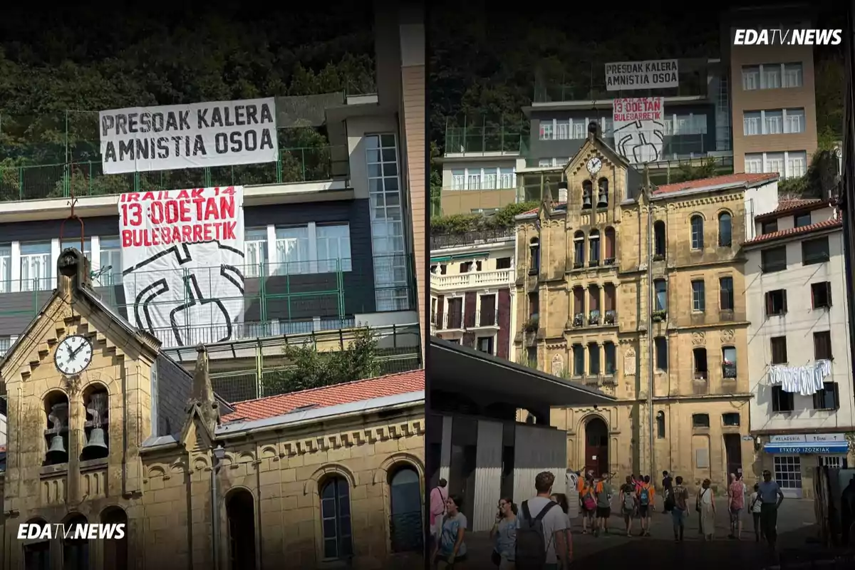 Dos pancartas grandes con mensajes en euskera cuelgan de un edificio moderno detrás de una iglesia antigua de piedra, mientras varias personas caminan por la plaza en un día soleado.