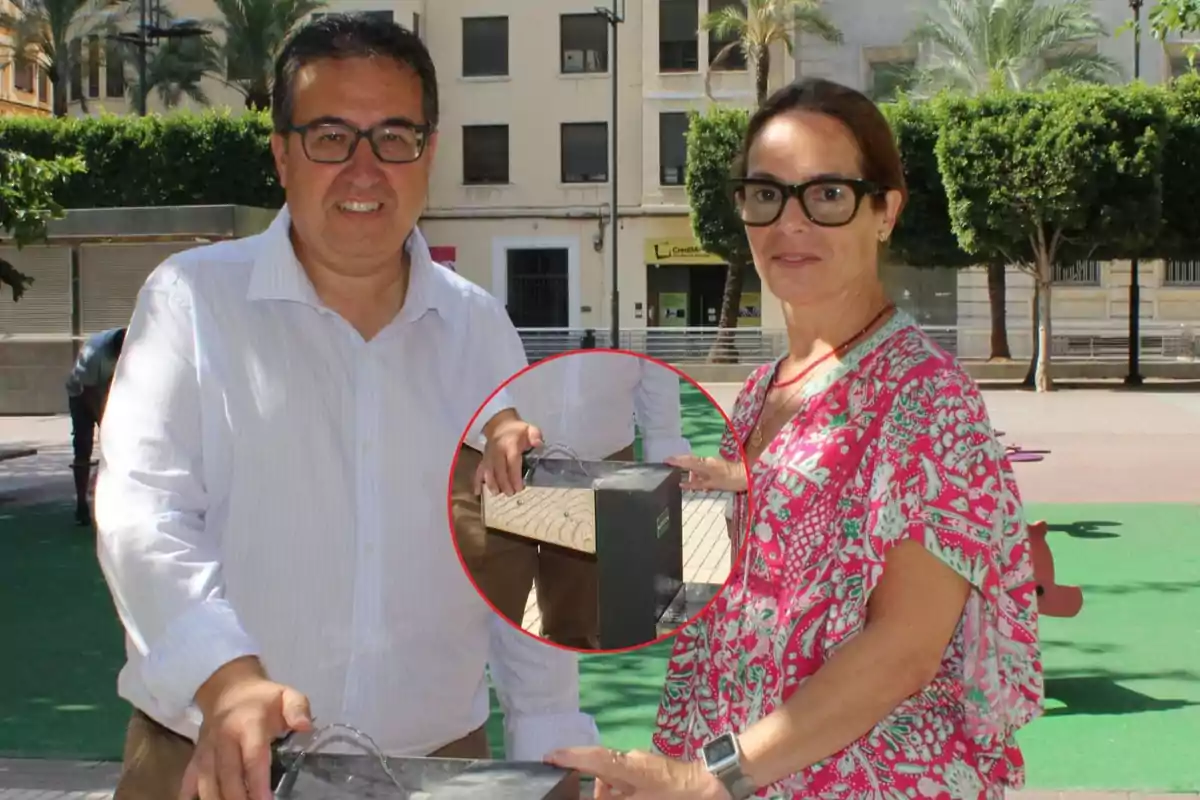 Dos personas posan en una plaza junto a una estructura de madera y metal con cables, mientras al fondo se observan árboles y edificios
