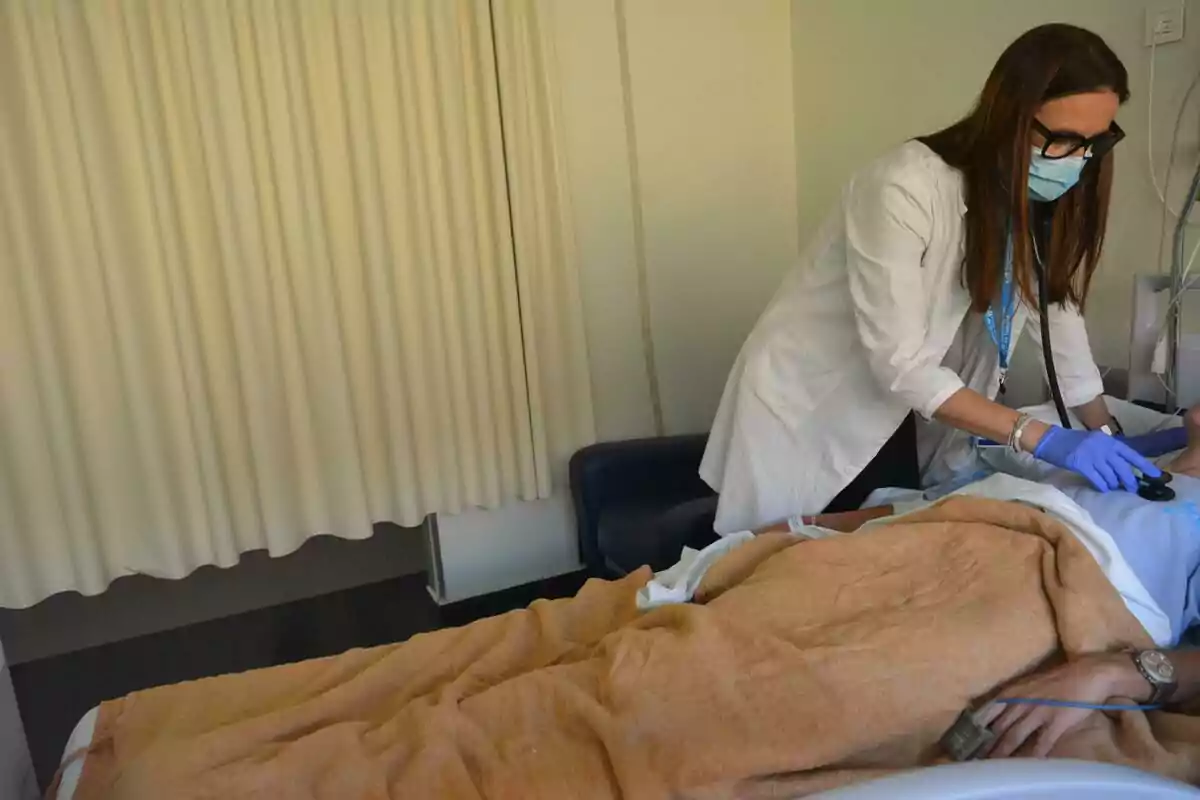 Doctora con bata blanca y mascarilla revisando a un paciente acostado en una cama de hospital cubierto con una manta marrón