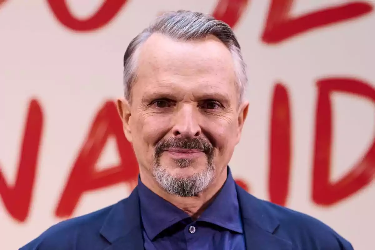 Hombre de cabello canoso y barba corta con saco azul posando frente a un fondo claro con letras rojas desenfocadas Hombre de cabello canoso y barba corta con saco azul posando frente a un fondo claro con letras rojas desenfocadas