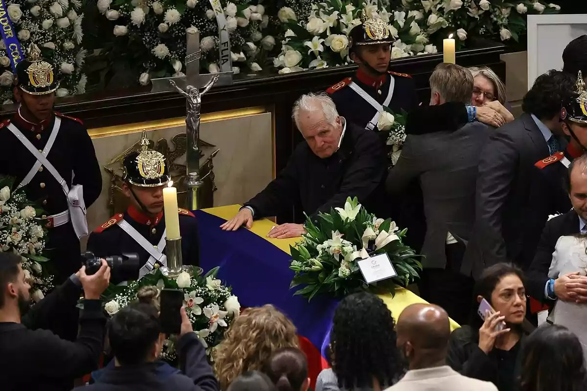 Un hombre mayor apoya sus manos sobre un ataúd cubierto con la bandera de Colombia, rodeado de flores blancas, guardias de uniforme y personas que se abrazan en un ambiente solemne de funeral. Un hombre mayor apoya sus manos sobre un ataúd cubierto con la bandera de Colombia, rodeado de flores blancas, guardias de uniforme y personas que se abrazan en un ambiente solemne de funeral.