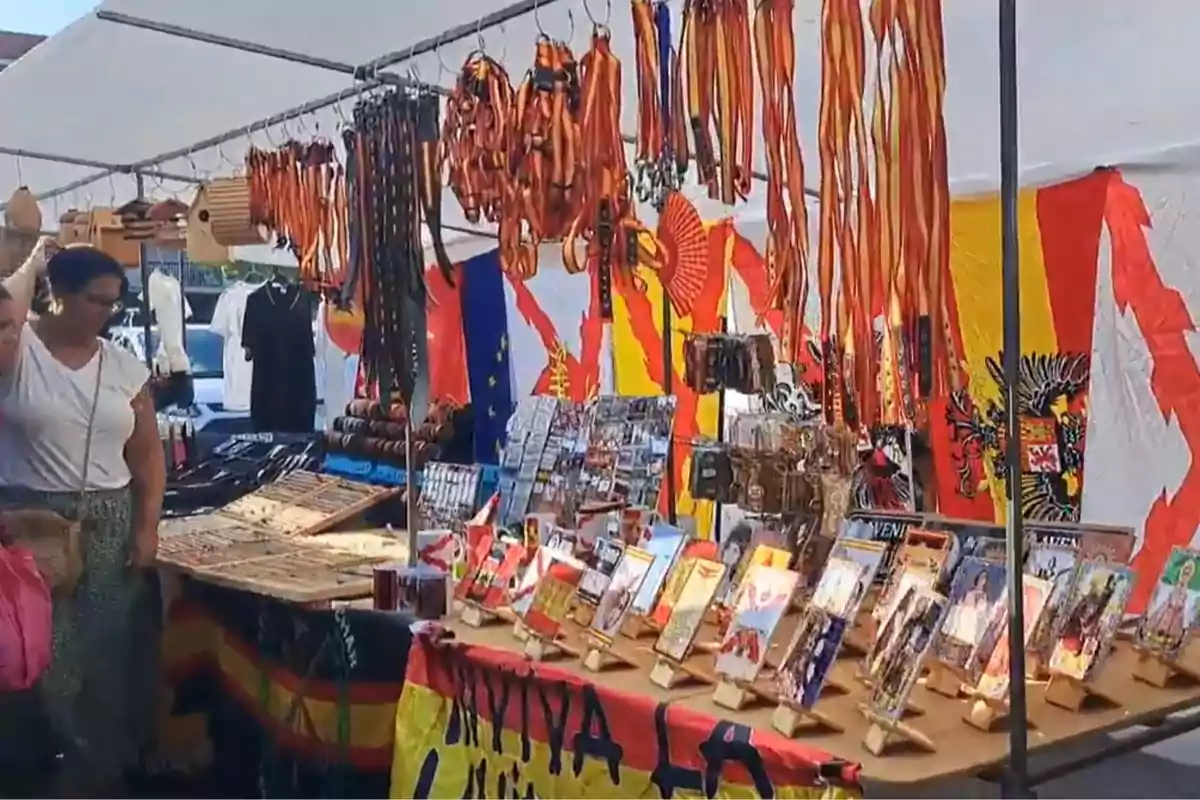Puesto de mercado al aire libre con cintas, pulseras, llaveros y postales decorativas, atendido por una mujer y decorado con banderas de colores vivos Puesto de mercado al aire libre con cintas, pulseras, llaveros y postales decorativas, atendido por una mujer y decorado con banderas de colores vivos