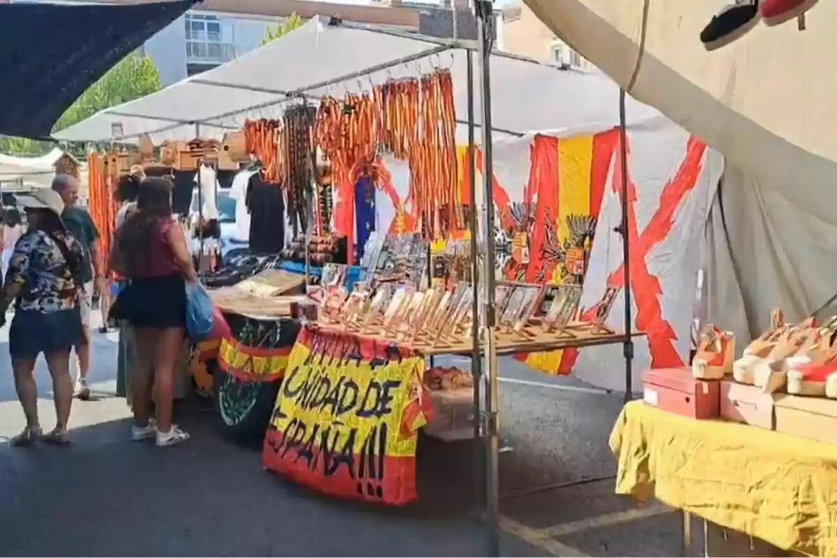 Puesto de mercado al aire libre con banderas de España, cintas rojigualdas, recuerdos y un cartel que dice defensa unidad de España, mientras varias personas observan los productos expuestos.