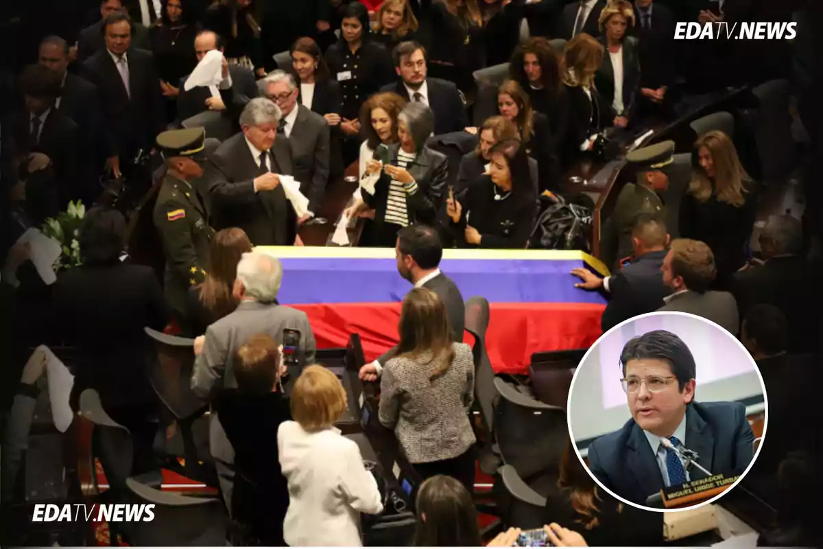Personas reunidas alrededor de un ataúd cubierto con la bandera de Colombia en una ceremonia solemne, con un recuadro mostrando a un hombre de traje y gafas hablando en un micrófono