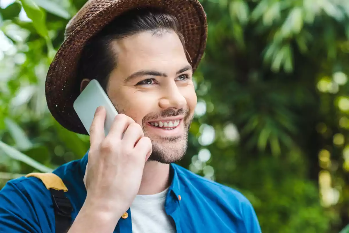 Hombre joven con sombrero sonríe mientras habla por teléfono móvil en un entorno natural