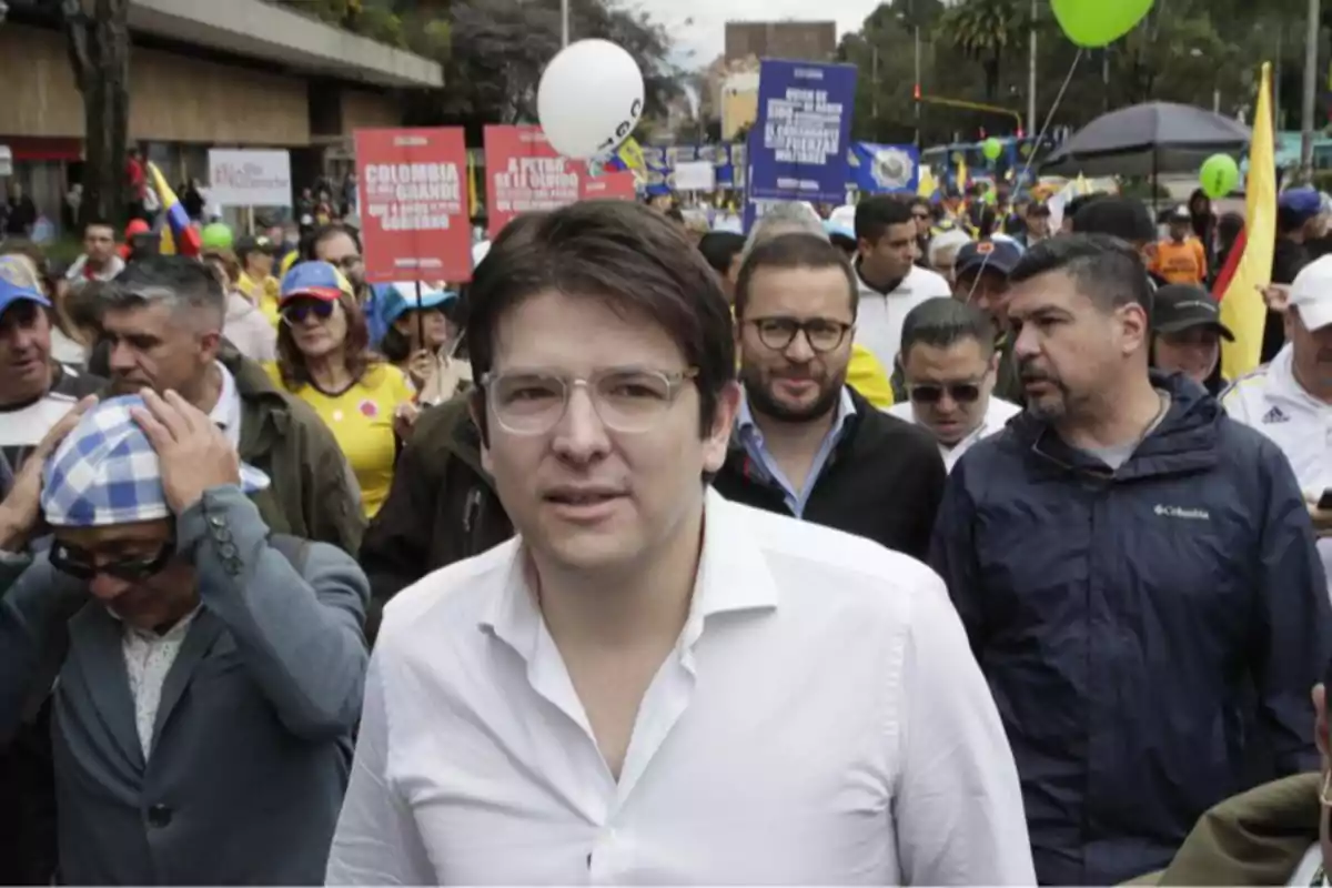 Un hombre con gafas y camisa blanca camina al frente de una multitud durante una manifestación en la calle, varias personas sostienen pancartas y globos de colores. Un hombre con gafas y camisa blanca camina al frente de una multitud durante una manifestación en la calle, varias personas sostienen pancartas y globos de colores.