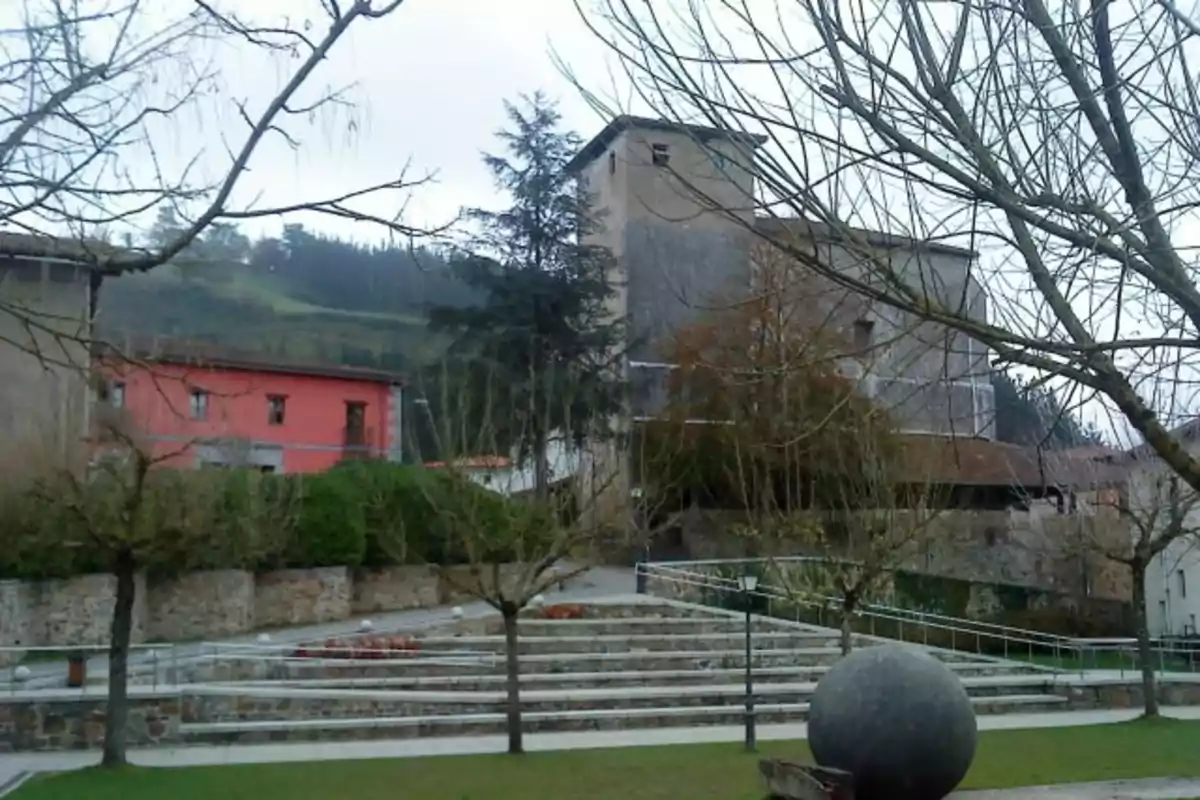 Plaza con escalinatas de piedra, árboles sin hojas, una escultura esférica y al fondo una torre antigua junto a una casa roja en un entorno rural Plaza con escalinatas de piedra, árboles sin hojas, una escultura esférica y al fondo una torre antigua junto a una casa roja en un entorno rural