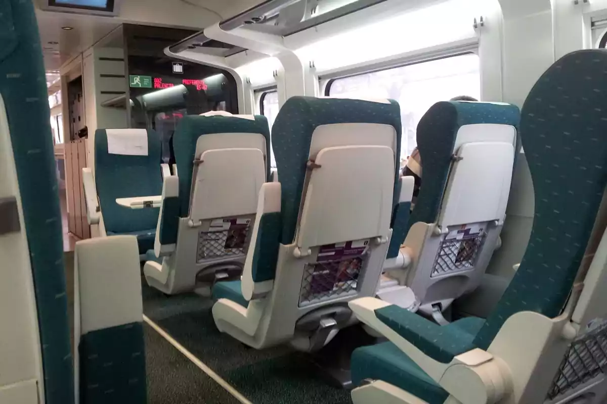 Interior de un tren moderno con asientos verdes y mesas plegables entre ellos, ventanas grandes y luz natural entrando