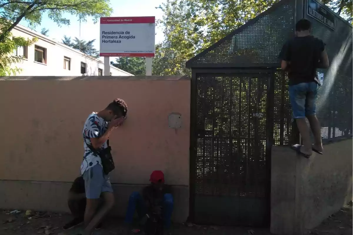 Tres jóvenes están junto a una pared y una reja metálica en la entrada de la Residencia de Primera Acogida Hortaleza en Madrid uno de ellos se cubre el rostro con las manos otro está sentado en el suelo y el tercero está de pie sobre el borde de la pared mirando hacia la reja