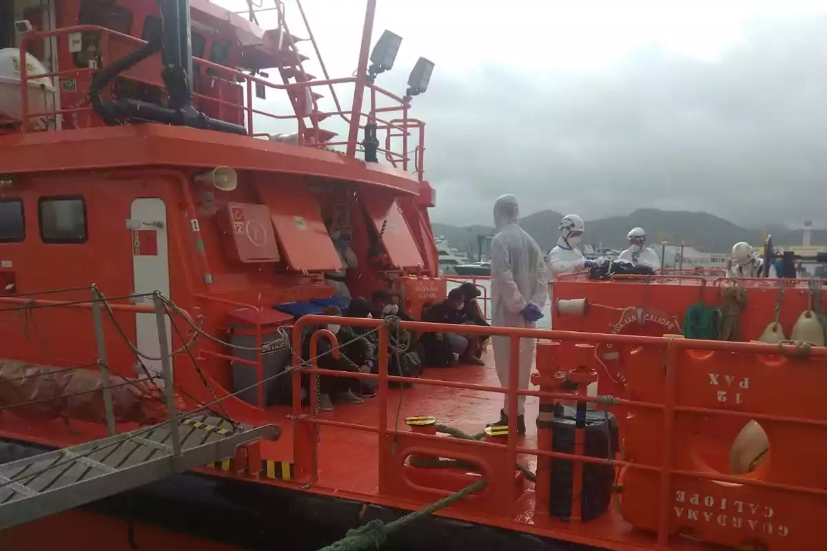 Personas sentadas y personal con trajes protectores en la cubierta de un barco de rescate naranja en un puerto bajo cielo nublado Personas sentadas y personal con trajes protectores en la cubierta de un barco de rescate naranja en un puerto bajo cielo nublado