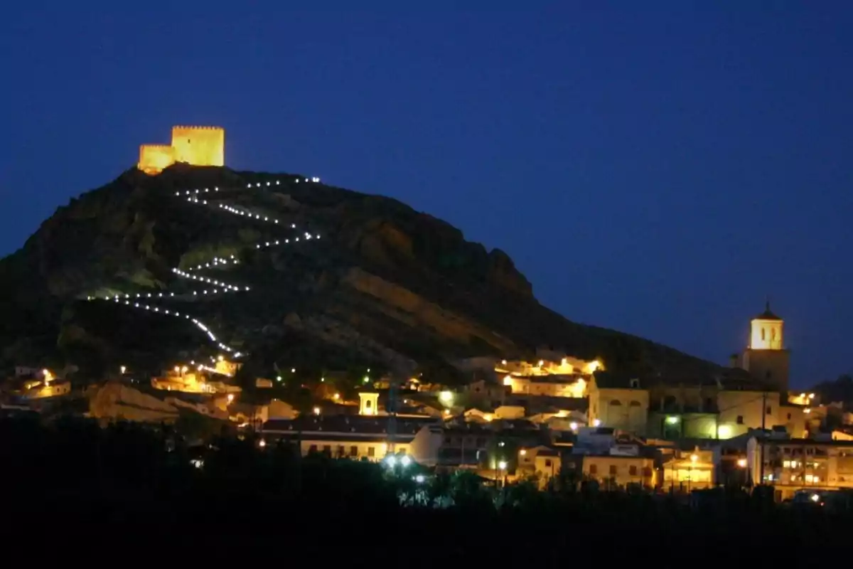 Vista nocturna de un castillo iluminado en lo alto de una colina con un sendero de luces en zigzag y un pueblo al pie de la montaña Vista nocturna de un castillo iluminado en lo alto de una colina con un sendero de luces en zigzag y un pueblo al pie de la montaña