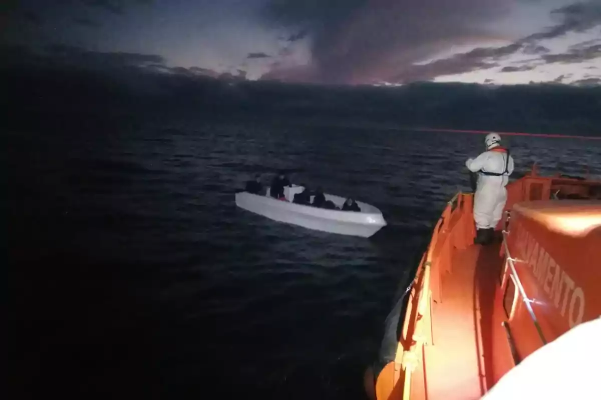 Un bote de rescate se acerca a una pequeña embarcación blanca con varias personas a bordo en el mar al anochecer