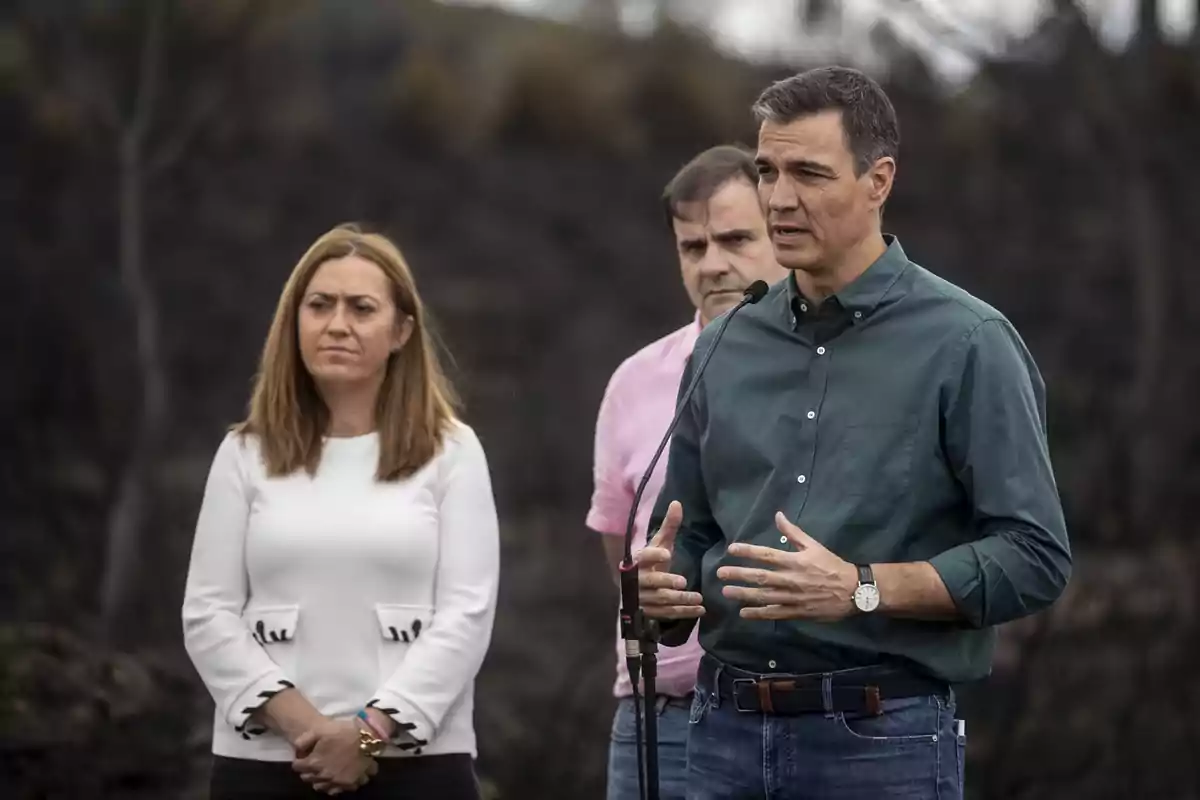 Tres personas de pie al aire libre con expresión seria mientras una de ellas habla frente a un micrófono Tres personas de pie al aire libre con expresión seria mientras una de ellas habla frente a un micrófono