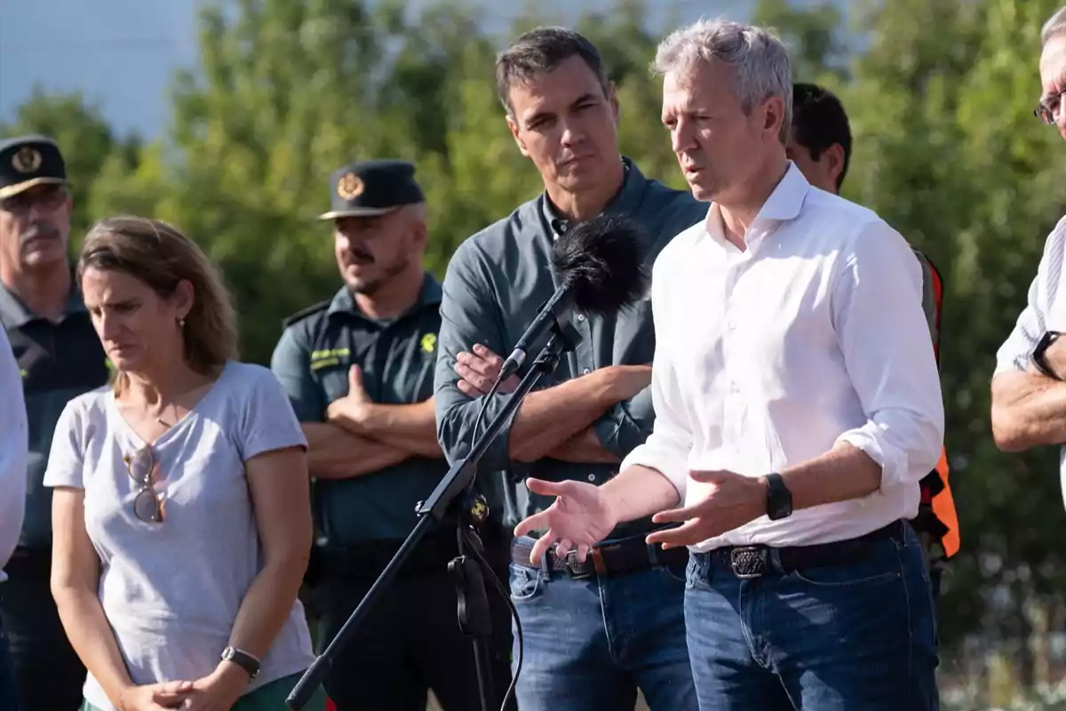 Un grupo de personas, incluyendo autoridades y agentes de seguridad, se encuentra reunido al aire libre mientras un hombre de cabello canoso habla frente a un micrófono Un grupo de personas, incluyendo autoridades y agentes de seguridad, se encuentra reunido al aire libre mientras un hombre de cabello canoso habla frente a un micrófono