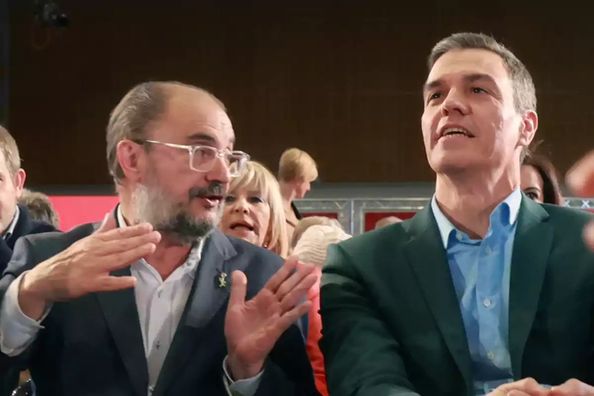 Dos hombres con traje conversan animadamente en un evento público rodeados de otras personas Dos hombres con traje conversan animadamente en un evento público rodeados de otras personas
