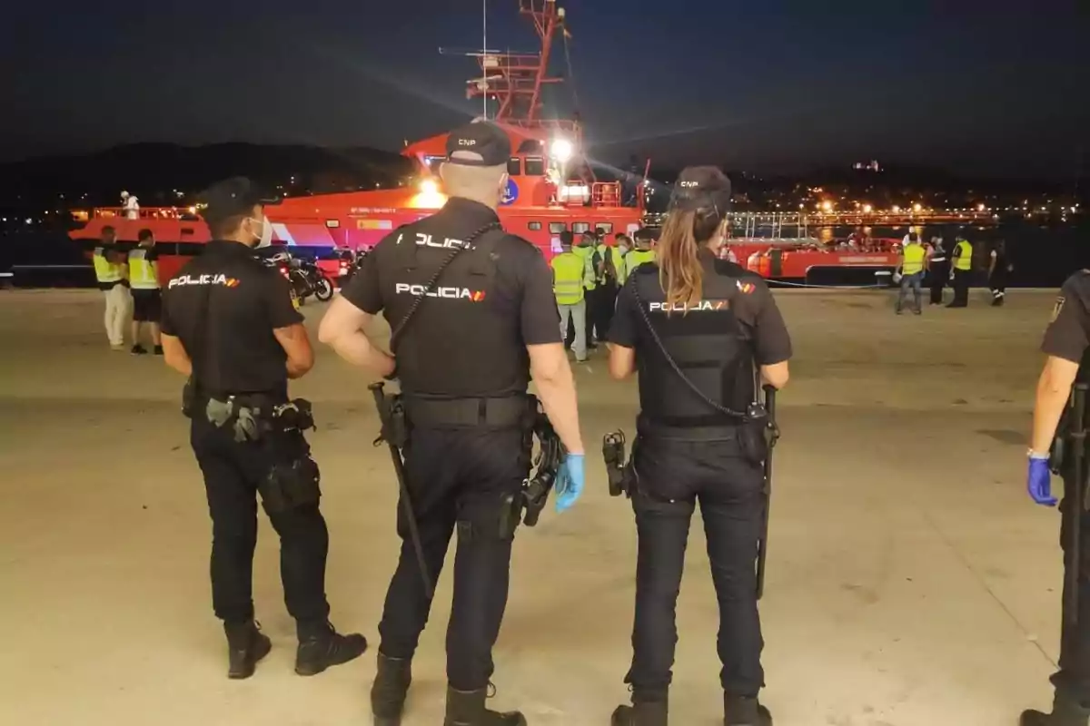 Varios policías de espaldas vigilan un muelle donde hay un barco de rescate iluminado y varias personas con chalecos reflectantes