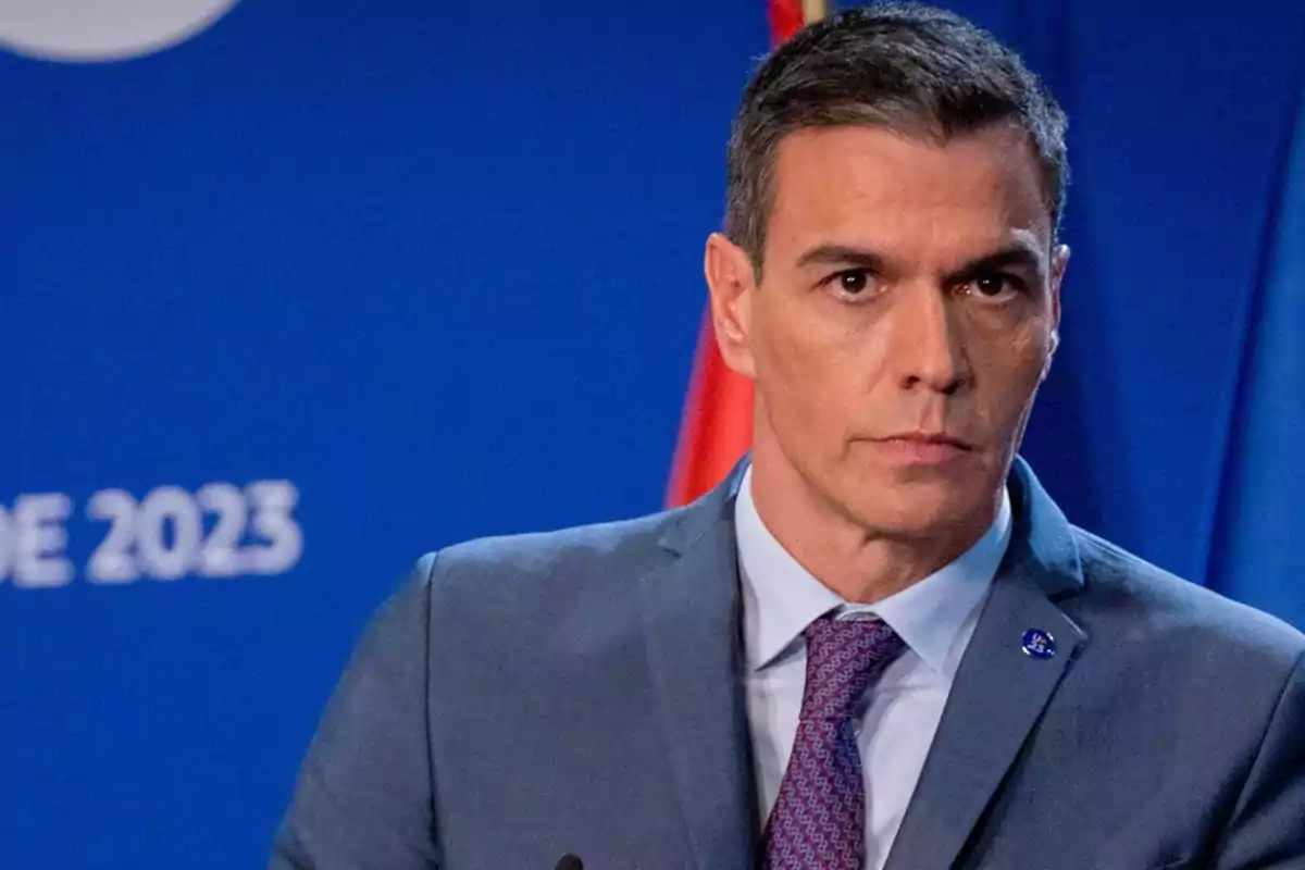 Hombre de cabello corto y traje gris con corbata morada frente a un fondo azul con letras blancas y una bandera roja detrás